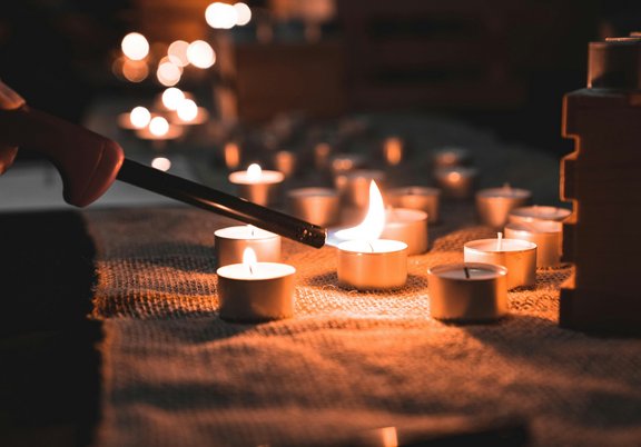 Das Bild zeigt eine Ablage, auf der einige Rechaudkerzen im Halbfinsteren stehen. Einige davon brennen. Eine Person zündet eine Kerze an.